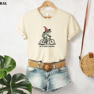 May include: Cream-colored t-shirt featuring a whimsical illustration of a green frog wearing a wizard hat and riding a bicycle. The text "OFF TO CAUSE A KERFUFFLE" is printed below the graphic. The shirt is paired with denim shorts and a woven belt.