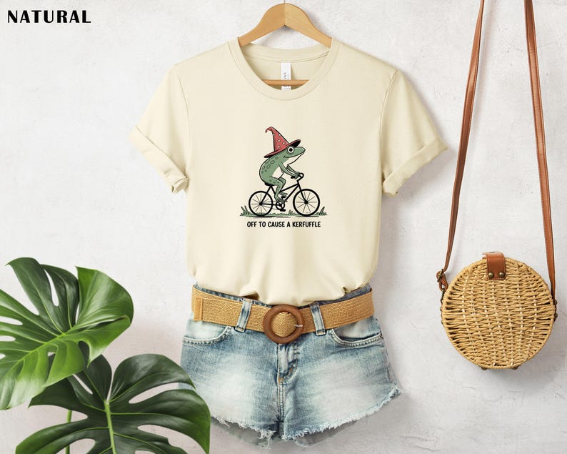 May include: Cream-colored t-shirt featuring a whimsical illustration of a green frog wearing a wizard hat and riding a bicycle. The text "OFF TO CAUSE A KERFUFFLE" is printed below the graphic. The shirt is paired with denim shorts and a woven belt.
