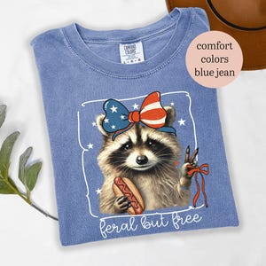 Op de afbeelding: Blauw Comfort Colors T-shirt met een wasbeer die een patriottische strik draagt en een hotdog vasthoudt. De wasbeer maakt het vredesteken. De tekst "feral but free" staat onderaan de afbeelding.