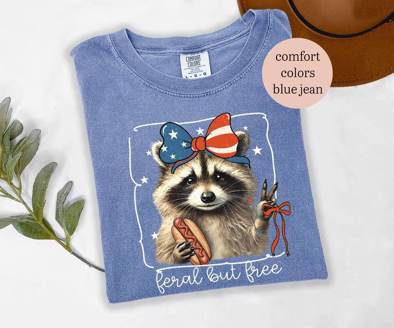 Op de afbeelding: Blauw Comfort Colors T-shirt met een wasbeer die een patriottische strik draagt en een hotdog vasthoudt. De wasbeer maakt het vredesteken. De tekst "feral but free" staat onderaan de afbeelding.