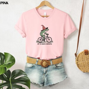 May include: A soft pink t-shirt with a whimsical graphic of a green frog riding a bicycle and wearing a witch hat. The text "OFF TO CAUSE A KERFUFFLE" is printed below the frog. The shirt is paired with denim shorts.