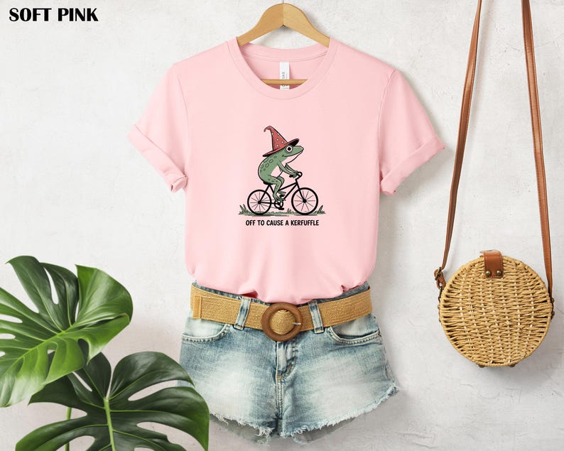 May include: A soft pink t-shirt with a whimsical graphic of a green frog riding a bicycle and wearing a witch hat. The text "OFF TO CAUSE A KERFUFFLE" is printed below the frog. The shirt is paired with denim shorts.