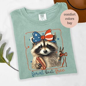 Op de afbeelding: Lichtgroen T-shirt met een wasbeer die een patriottische strik draagt en een hotdog vasthoudt. De wasbeer maakt het vredesteken. De tekst "feral but free" staat onderaan de afbeelding.