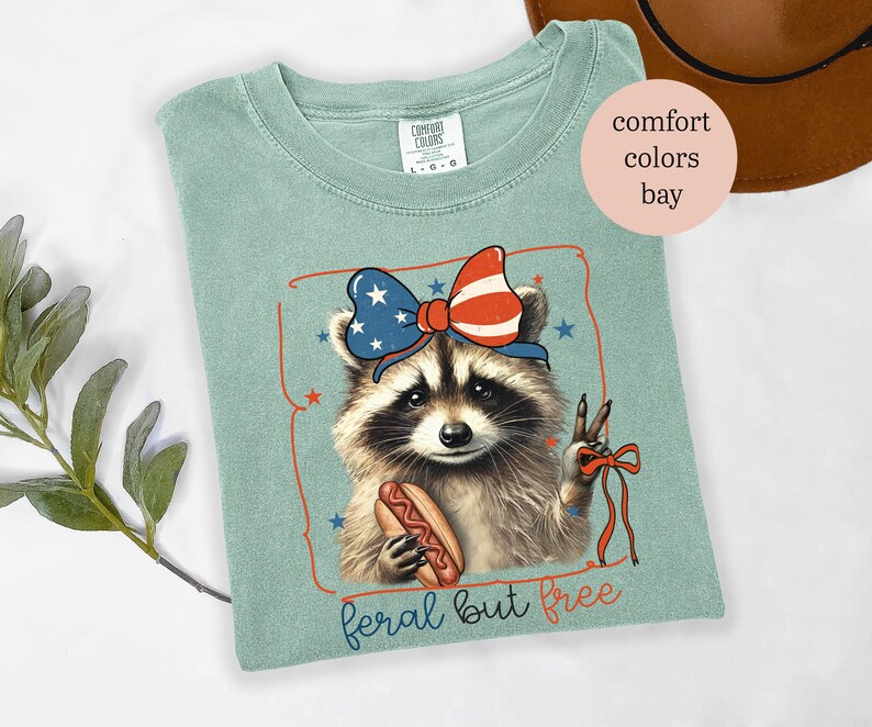 Op de afbeelding: Lichtgroen T-shirt met een wasbeer die een patriottische strik draagt en een hotdog vasthoudt. De wasbeer maakt het vredesteken. De tekst "feral but free" staat onderaan de afbeelding.