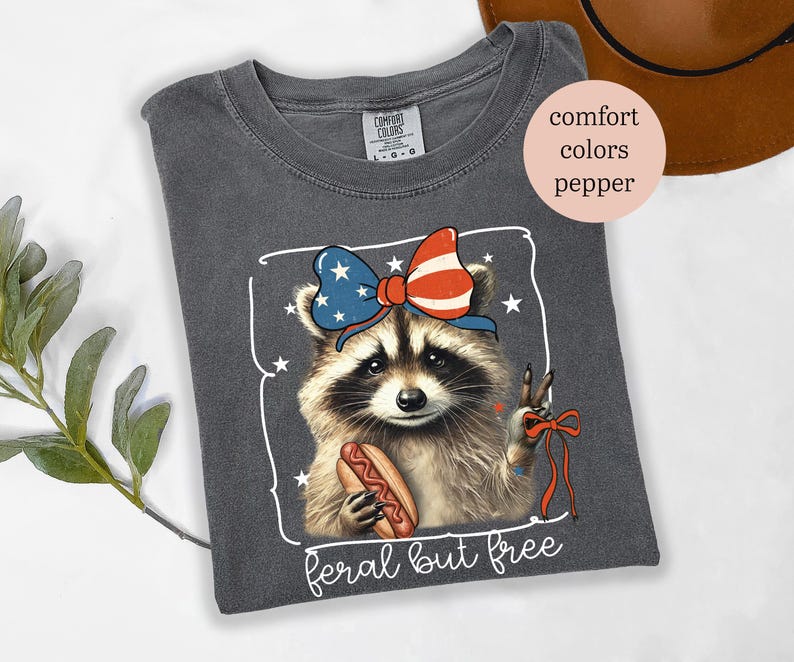 Op de afbeelding: Donkergrijs T-shirt met een wasbeer die een patriottische strik draagt en een hotdog vasthoudt. De tekst "feral but free" staat onder de afbeelding van de wasbeer.