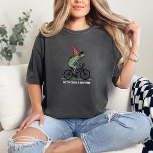 May include: Dark gray t-shirt featuring a green frog wearing a witch hat and riding a bicycle. The text "OFF TO CAUSE A KERFUFFLE" is printed below the graphic. The shirt is a casual, unisex style.