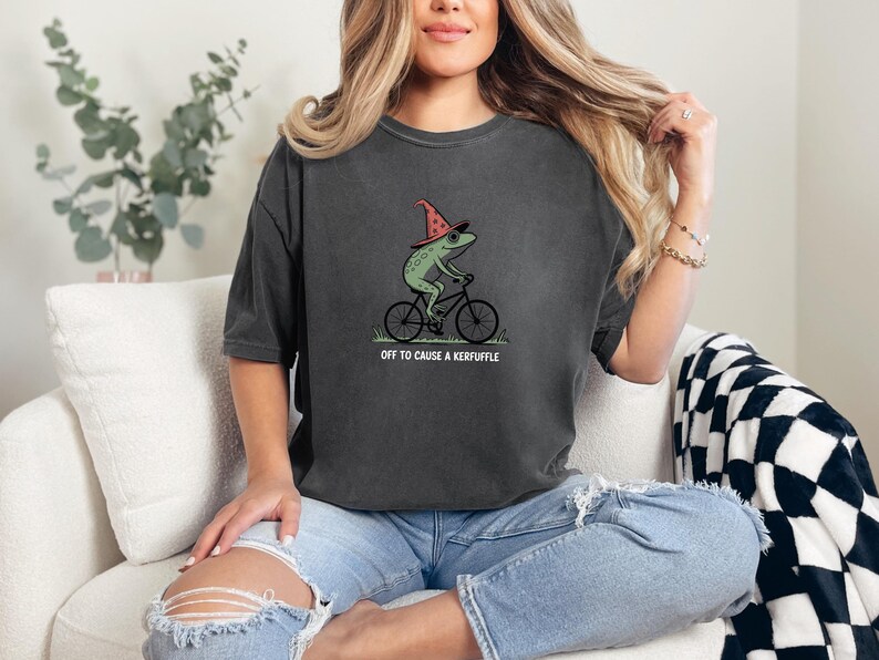 May include: Dark gray t-shirt featuring a green frog wearing a witch hat and riding a bicycle. The text "OFF TO CAUSE A KERFUFFLE" is printed below the graphic. The shirt is a casual, unisex style.