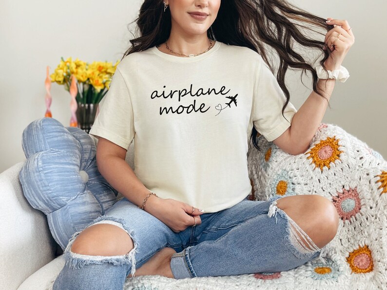 K&ouml;nnte beinhalten: Eine Frau tr&auml;gt ein cremefarbenes T-Shirt mit dem Text "airplane mode" und einer kleinen Flugzeuggrafik. Sie sitzt auf einem Sofa mit einem blauen Kissen und einer geh&auml;kelten Decke.
