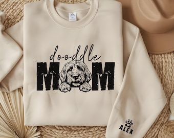 May include: A beige sweatshirt with the text "doodle MOM" in black with a doodle dog peeking out from behind the letters. The sweatshirt has a black paw print with the name "ALEX" on the sleeve.