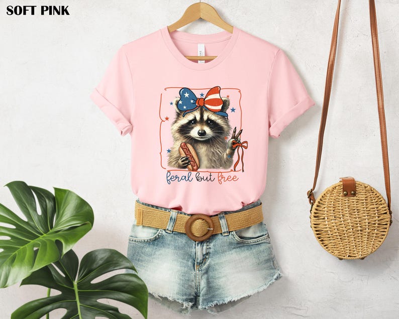 Op de afbeelding: Zachtroze T-shirt met een wasbeer die een patriottische strik draagt en een hotdog vasthoudt. De wasbeerillustratie is omlijst met sterren en de woorden "feral but free". Het T-shirt wordt op een hanger getoond.