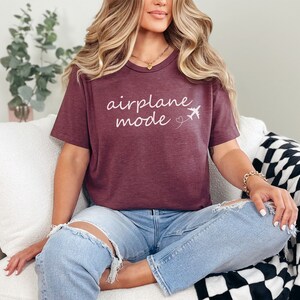 K&ouml;nnte beinhalten: Ein maroonfarbenes T-Shirt mit dem Text "airplane mode" in wei&szlig;er Kursivschrift und einer kleinen Flugzeuggrafik. Das Shirt wird von einer Person getragen, die auf einem wei&szlig;en Sofa sitzt.