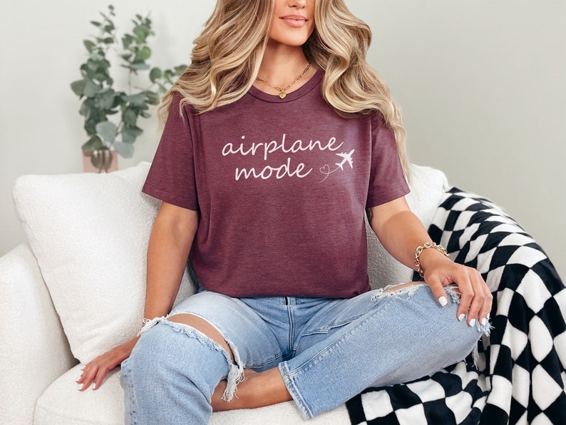 K&ouml;nnte beinhalten: Ein maroonfarbenes T-Shirt mit dem Text "airplane mode" in wei&szlig;er Kursivschrift und einer kleinen Flugzeuggrafik. Das Shirt wird von einer Person getragen, die auf einem wei&szlig;en Sofa sitzt.