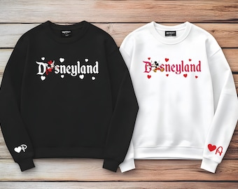 May include: A black sweatshirt with a white Disneyland logo featuring Minnie Mouse and white hearts, and a white sweatshirt with a red Disneyland logo featuring Mickey Mouse and red hearts. Both feature small heart and letter graphics on the cuffs.