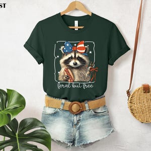 Op de afbeelding: Donkergroen t-shirt met een wasbeer met een patriottische strik en een hotdog. De wasbeer staat in een wit kader met de tekst "feral but free" eronder. Het t-shirt hangt aan een houten hanger.