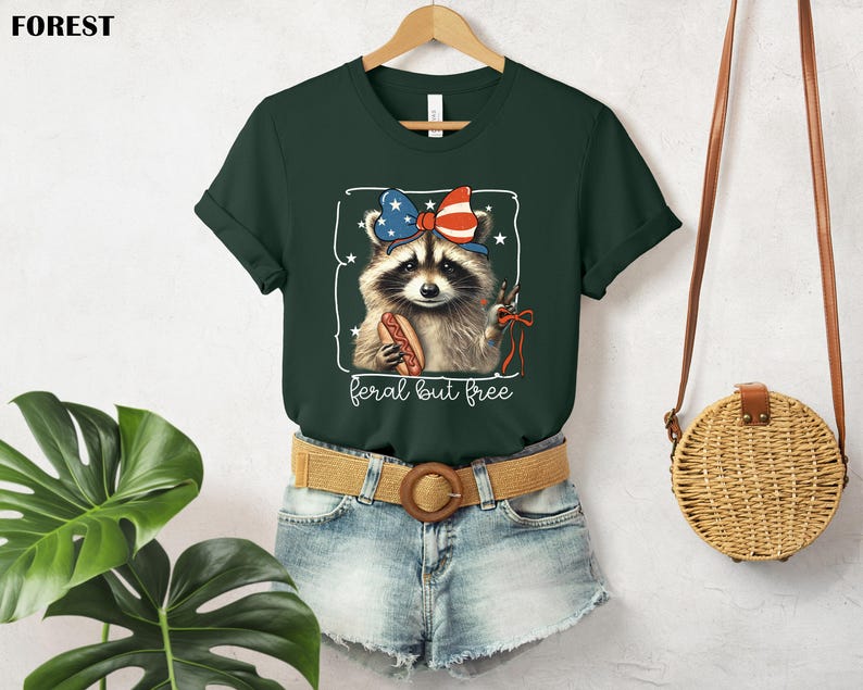 Op de afbeelding: Donkergroen t-shirt met een wasbeer met een patriottische strik en een hotdog. De wasbeer staat in een wit kader met de tekst "feral but free" eronder. Het t-shirt hangt aan een houten hanger.