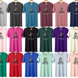 May include: A collection of short-sleeved t-shirts in various colors, each featuring a cartoon frog riding a bicycle and wearing a hat. The text "TRY TO CATCH A BUBBLE" is printed below the frog. The shirts are displayed on wooden hangers.