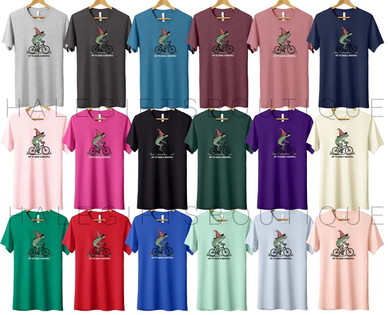 May include: A collection of short-sleeved t-shirts in various colors, each featuring a cartoon frog riding a bicycle and wearing a hat. The text "TRY TO CATCH A BUBBLE" is printed below the frog. The shirts are displayed on wooden hangers.