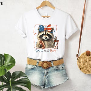 Op de afbeelding: Wit T-shirt met een wasbeer die een patriottische strik draagt en een hotdog vasthoudt. De wasbeer staat in een rode omlijsting met sterren. De tekst "feral but free" staat eronder.
