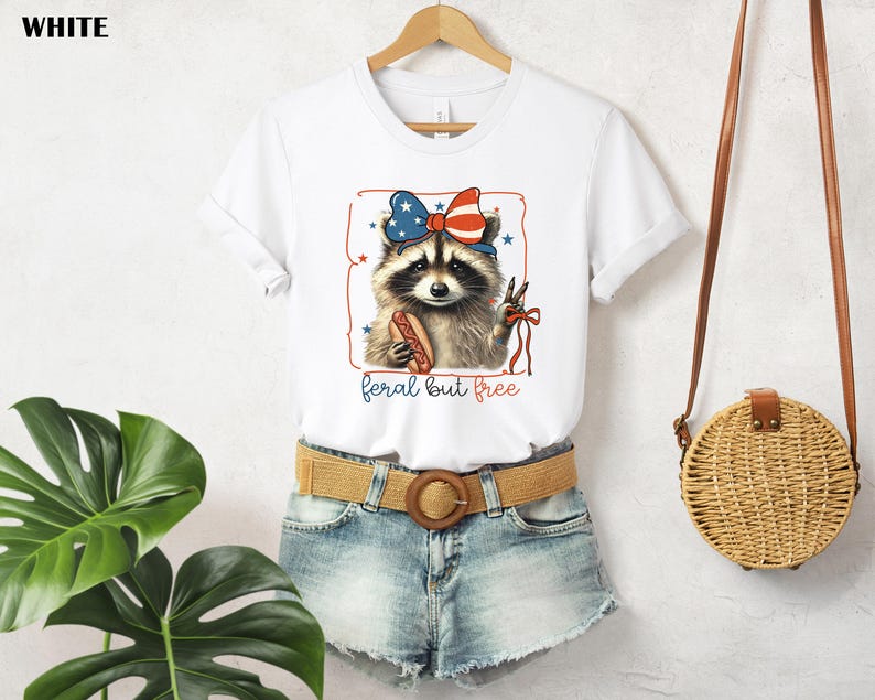 Op de afbeelding: Wit T-shirt met een wasbeer die een patriottische strik draagt en een hotdog vasthoudt. De wasbeer staat in een rode omlijsting met sterren. De tekst "feral but free" staat eronder.