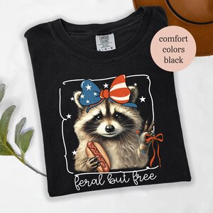 Op de afbeelding: Zwart T-shirt met een afbeelding van een wasbeer met een patriottische strik en een hotdog. De tekst "feral but free" staat onder de afbeelding van de wasbeer.