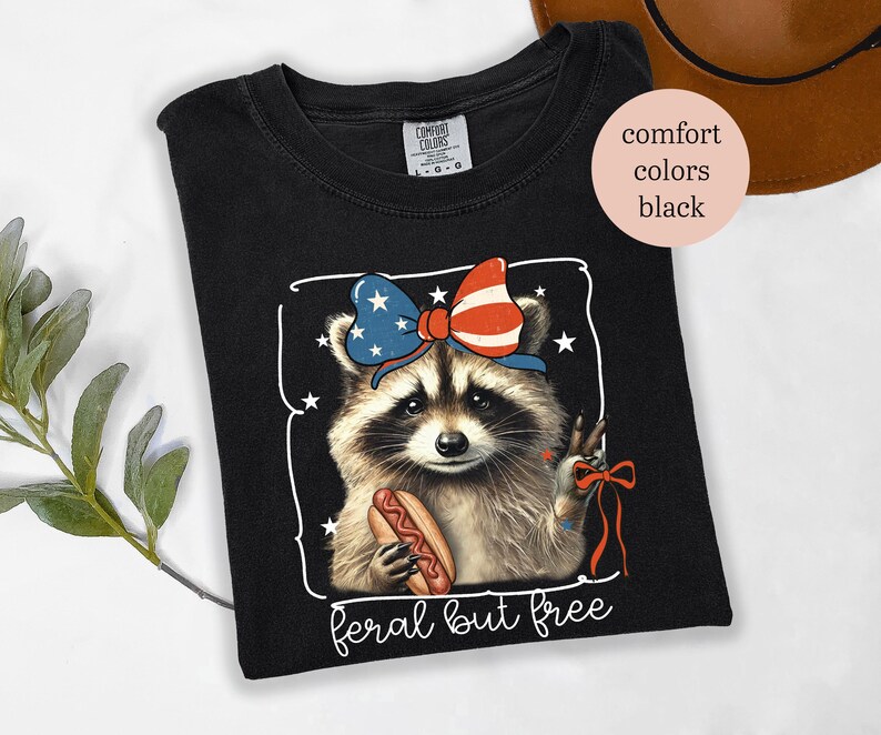 Op de afbeelding: Zwart T-shirt met een afbeelding van een wasbeer met een patriottische strik en een hotdog. De tekst "feral but free" staat onder de afbeelding van de wasbeer.