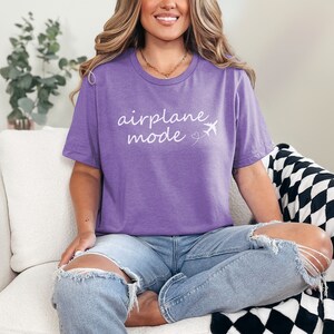 K&ouml;nnte beinhalten: Eine Frau tr&auml;gt ein lila T-Shirt mit dem Text "airplane mode" in wei&szlig;en Buchstaben und einer kleinen Flugzeuggrafik. Sie sitzt auf einem wei&szlig;en Sofa mit einer schwarz-wei&szlig;en karierten Wolldecke.