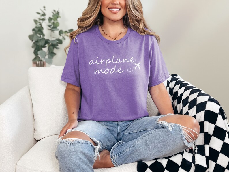 K&ouml;nnte beinhalten: Eine Frau tr&auml;gt ein lila T-Shirt mit dem Text "airplane mode" in wei&szlig;en Buchstaben und einer kleinen Flugzeuggrafik. Sie sitzt auf einem wei&szlig;en Sofa mit einer schwarz-wei&szlig;en karierten Wolldecke.