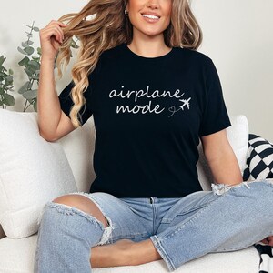 K&ouml;nnte beinhalten: Eine Frau tr&auml;gt ein schwarzes T-Shirt mit dem Text "airplane mode" und einer kleinen Flugzeuggrafik. Sie sitzt auf einem wei&szlig;en Sofa mit einer schwarz-wei&szlig;en karierten Wolldecke.