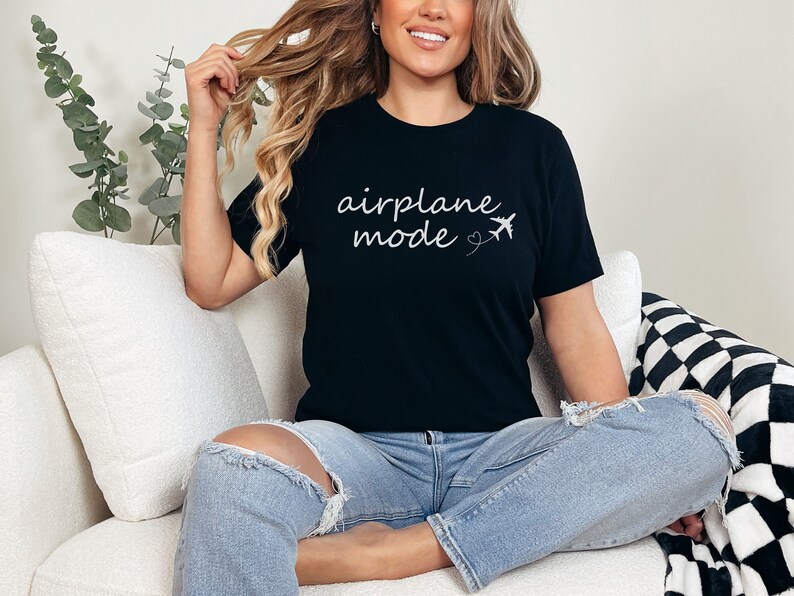 K&ouml;nnte beinhalten: Eine Frau tr&auml;gt ein schwarzes T-Shirt mit dem Text "airplane mode" und einer kleinen Flugzeuggrafik. Sie sitzt auf einem wei&szlig;en Sofa mit einer schwarz-wei&szlig;en karierten Wolldecke.
