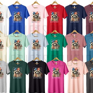 Op de afbeelding: Een verzameling korte mouwen T-shirts in verschillende kleuren, elk met een wasbeer-afbeelding met een Amerikaanse vlag strik en de tekst "Born but Free". De shirts worden op houten hangers getoond.