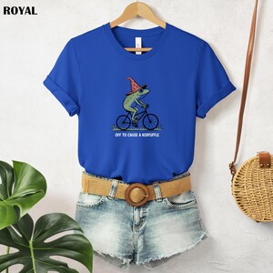 May include: A royal blue t-shirt featuring a whimsical illustration of a green frog wearing a wizard hat and riding a bicycle. The text "OFF TO CAUSE A KERFUFFLE" is below the frog. The shirt is hanging on a wooden hanger.