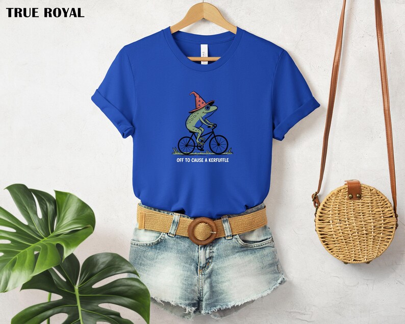 May include: A royal blue t-shirt featuring a whimsical illustration of a green frog wearing a wizard hat and riding a bicycle. The text "OFF TO CAUSE A KERFUFFLE" is below the frog. The shirt is hanging on a wooden hanger.