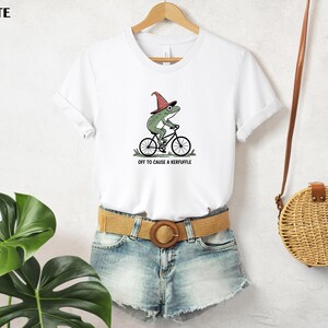 May include: White t-shirt featuring a green frog wearing a witch hat and riding a bicycle. The text "off to cause a kerfuffle" is printed below the graphic. The shirt is hanging on a wooden hanger.