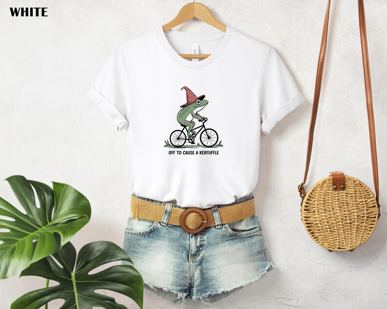 May include: White t-shirt featuring a green frog wearing a witch hat and riding a bicycle. The text "off to cause a kerfuffle" is printed below the graphic. The shirt is hanging on a wooden hanger.