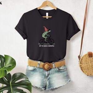 May include: Black t-shirt featuring a green frog riding a bicycle and wearing a red wizard hat. The text "OFF TO CAUSE A KERFUFFLE" is printed below the frog. The shirt is hanging on a wooden hanger.