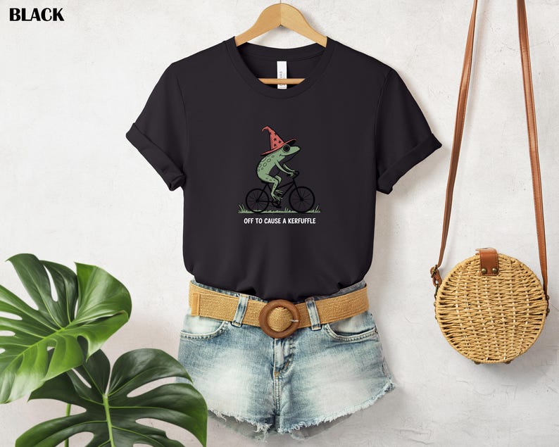 May include: Black t-shirt featuring a green frog riding a bicycle and wearing a red wizard hat. The text "OFF TO CAUSE A KERFUFFLE" is printed below the frog. The shirt is hanging on a wooden hanger.