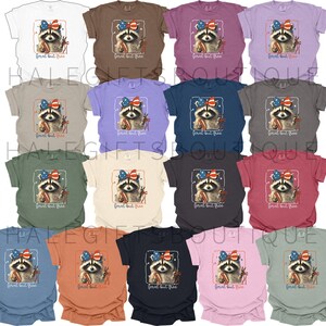 Op de afbeelding: Een collectie t-shirts in diverse kleuren, elk met een wasbeer met een patriottische strik en vuurwerk. De tekst "feral but free" staat eronder. De shirts zijn verkrijgbaar in verschillende kleuren.