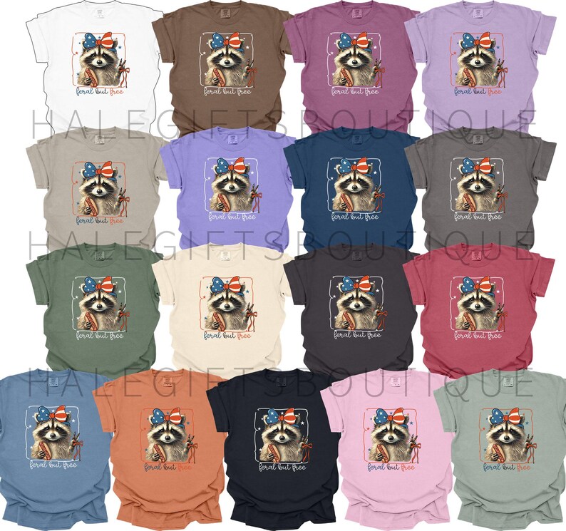 Op de afbeelding: Een collectie t-shirts in diverse kleuren, elk met een wasbeer met een patriottische strik en vuurwerk. De tekst "feral but free" staat eronder. De shirts zijn verkrijgbaar in verschillende kleuren.