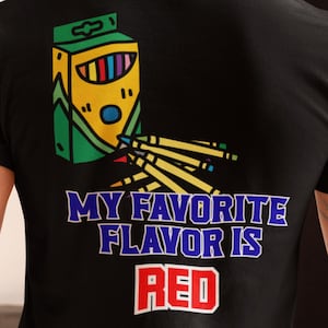 May include: Black t-shirt featuring a crayon box design with the text "MY FAVORITE FLAVOR IS RED." The graphic includes yellow, green, blue, and red colours. The shirt is made of a soft material, perfect for casual wear. The design is eye-catching.