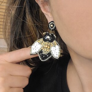 May include: A pair of dangle earrings featuring a black, gold, and white beaded bee design. The bee has a black body with gold accents and white wings.