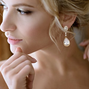 May include: Gold-toned drop earrings with clear, sparkling crystals. The earrings feature an oval top, three small crystals, and a large, pear-shaped crystal. The design is elegant and suitable for special occasions.