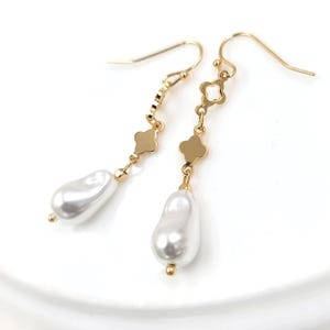 May include: A pair of gold-colored dangle earrings featuring white, teardrop-shaped faux pearls. The earrings have a delicate design with clover-shaped accents and a hook closure. The pearls have a lustrous, pearlescent finish, adding a touch of elegance.