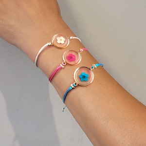 May include: Three bracelets with different colored cords and round charms featuring pressed flowers. The charms are gold-toned and have a clear glass or plastic cover. The flowers are white, pink, and blue.