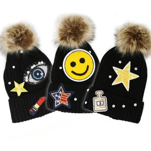 May include: Three black knit beanies with faux fur pom-poms. Each hat features decorative patches, including a yellow star, a lipstick, a smiley face, an eye, a star with a US flag design, and a perfume bottle. The hats are embellished with small, round, white rhinestones.