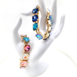 May include: Two gold bracelets with colorful square-shaped crystals. The bracelet on the left has pink, white, and blue crystals. The bracelet on the right has blue, teal, and iridescent crystals.
