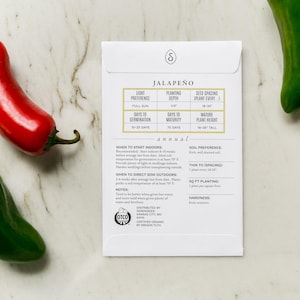May include: A white seed packet labeled "Jalape&ntilde;o" with planting instructions, accompanied by a red and two green jalape&ntilde;o peppers. The packet includes details on light preference, planting depth, and plant height.