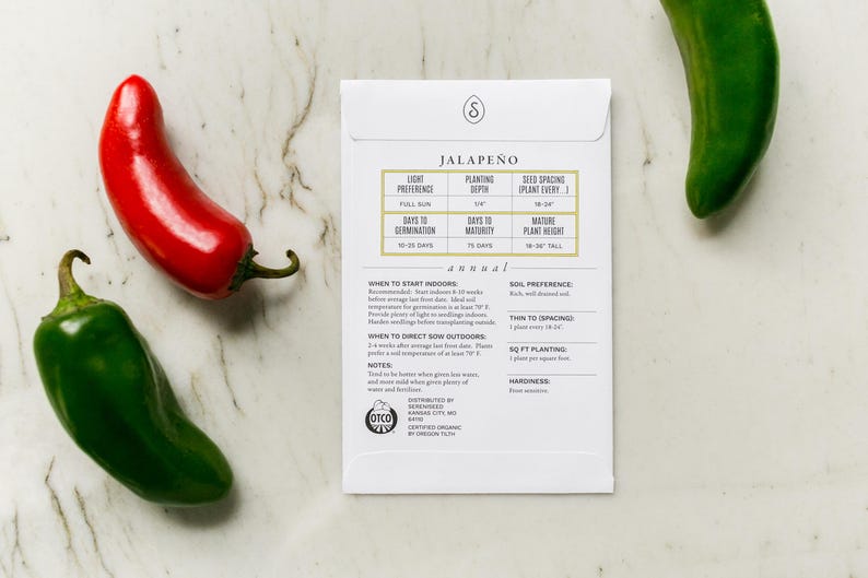 May include: A white seed packet labeled "Jalape&ntilde;o" with planting instructions, accompanied by a red and two green jalape&ntilde;o peppers. The packet includes details on light preference, planting depth, and plant height.