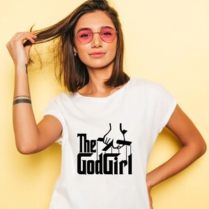 May include: White t-shirt with the text "The GodGirl" in a black, stylized font. The design includes a hand holding puppet strings. The shirt is short-sleeved and has a round neckline.