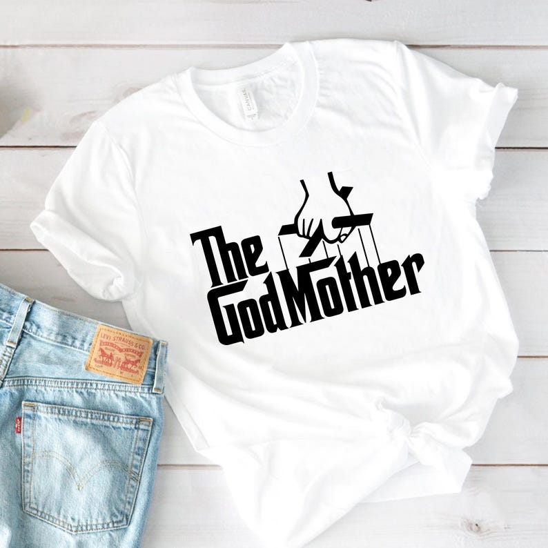 May include: White t-shirt with the text "The GodMother" in a black, stylized font, featuring a puppet master design. The shirt is paired with light blue denim jeans.