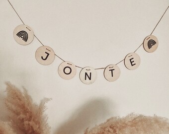 Peut inclure: Une bannière en bois avec les lettres "J O N T E" en lettres noires sur un fond beige. La bannière est enfilée sur une ficelle et a un petit motif arc-en-ciel à chaque extrémité.